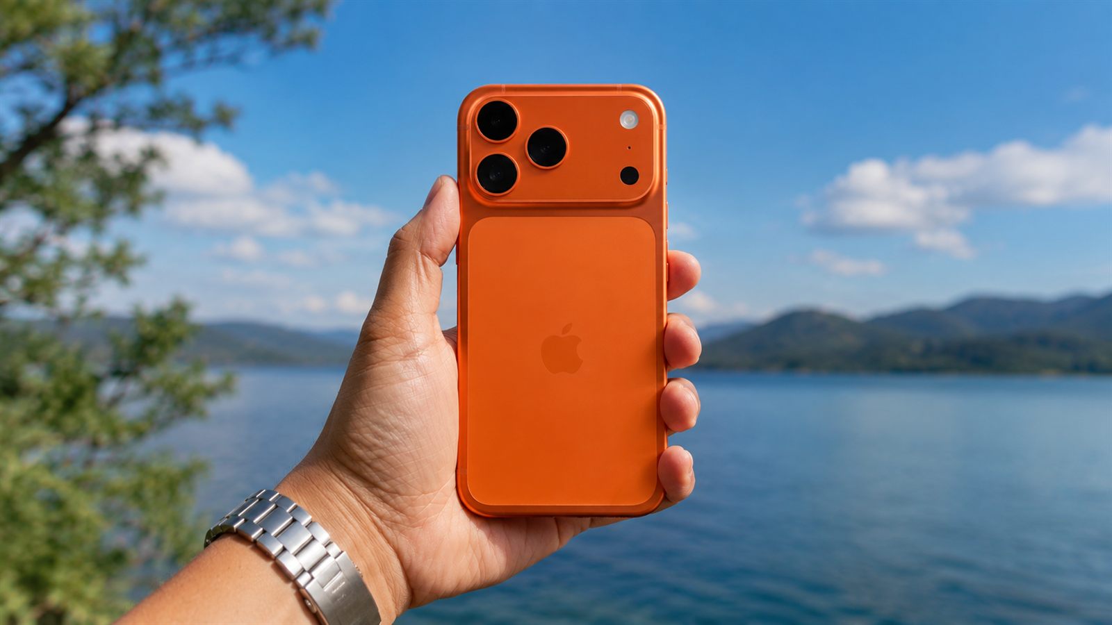 A hand holding an iPhone 17 Pro Max in Cosmic Orange outdoors by a lake, showing its triple-lens rear camera design